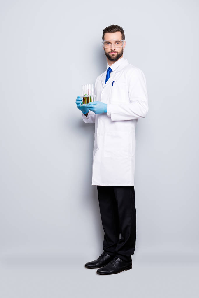 Volledige grootte volledige portret van aantrekkelijke stijlvolle wetenschapper in witte laboratoriumjas, zwarte broek, schoenen, gelijkspel bedrijf reageerbuisjes met multi-gekleurde vloeistof, kijken camera, geïsoleerd op een grijze achtergrond - Foto, afbeelding
