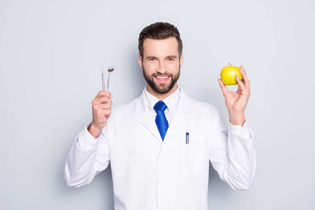 Portrait of successful toothy dentist in white lab coat, blue tie, holding equipments and green apple in hands, isolated on grey background, recommend taking care about teeth - Photo, Image