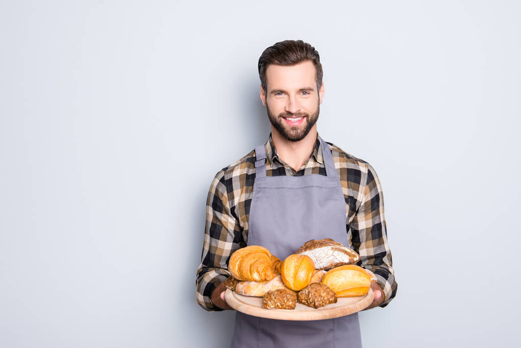 Portrait of friendly attractive baker in uniform with stubble having present showing tray with bakery products, looking at camera, isolated on grey background - Photo, Image