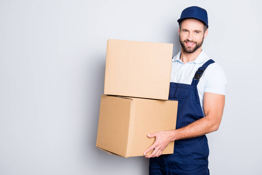 Portrait of attractive cheerful deliver in blue uniform with stubble looking at camera having two big boxes in hands, isolated on grey background - Photo, Image