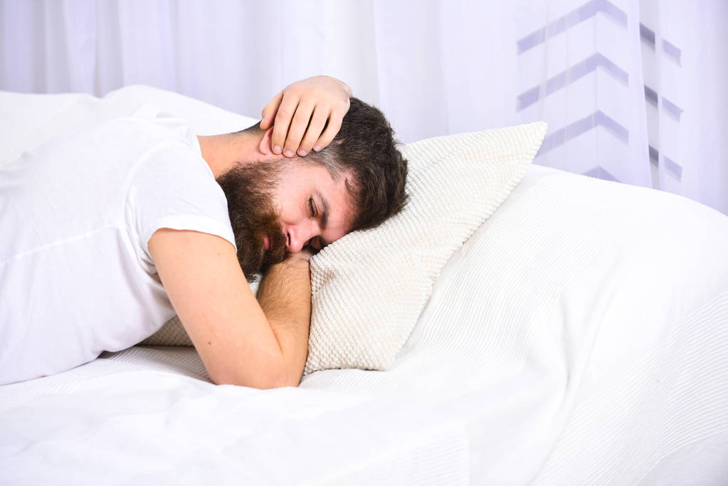 Mann im Hemd auf dem Bett liegend, Ohren mit Händen bedeckt, weißer Vorhang im Hintergrund. Weckruf-Konzept. Macho mit Bart und Schnurrbart weigert sich aufzustehen. Mann auf schläfrigem müden Gesicht schläft weiter - Foto, Bild