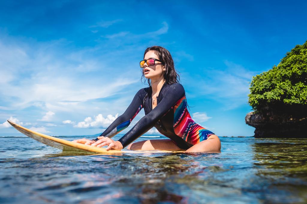 спортсменка в гидрокостюме и солнцезащитных очках на доске для серфинга в океане на пляже Нуса Дуа, Бали, Индонезия
 - Фото, изображение
