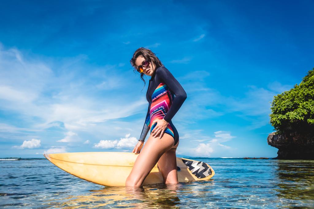 atraktivní mladá žena v obleku a sluneční brýle s Surf pózuje v oceánu na Nusa dua Beach, Bali, Indonésie - Fotografie, Obrázek
