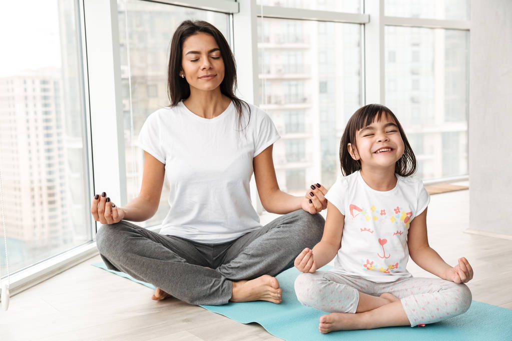 Mère paisible et petite fille méditant à la maison assis dans la pose du lotus sur le tapis et faisant geste de yoga mudra avec les doigts
 - Photo, image