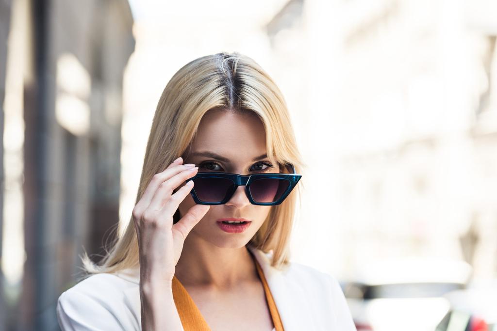 Porträt einer schönen jungen blonden Frau, die eine Sonnenbrille aufsetzt und in die Kamera blickt - Foto, Bild