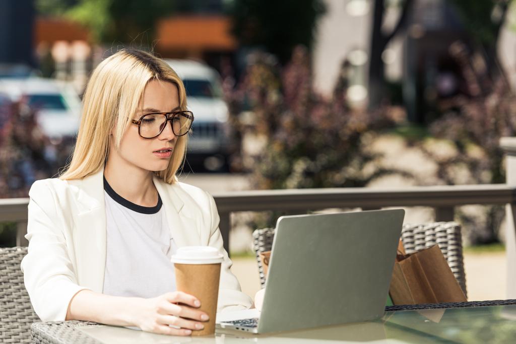 jonge blonde vrouw in brillen houden wegwerp koffiekopje en het gebruik van laptop - Foto, afbeelding