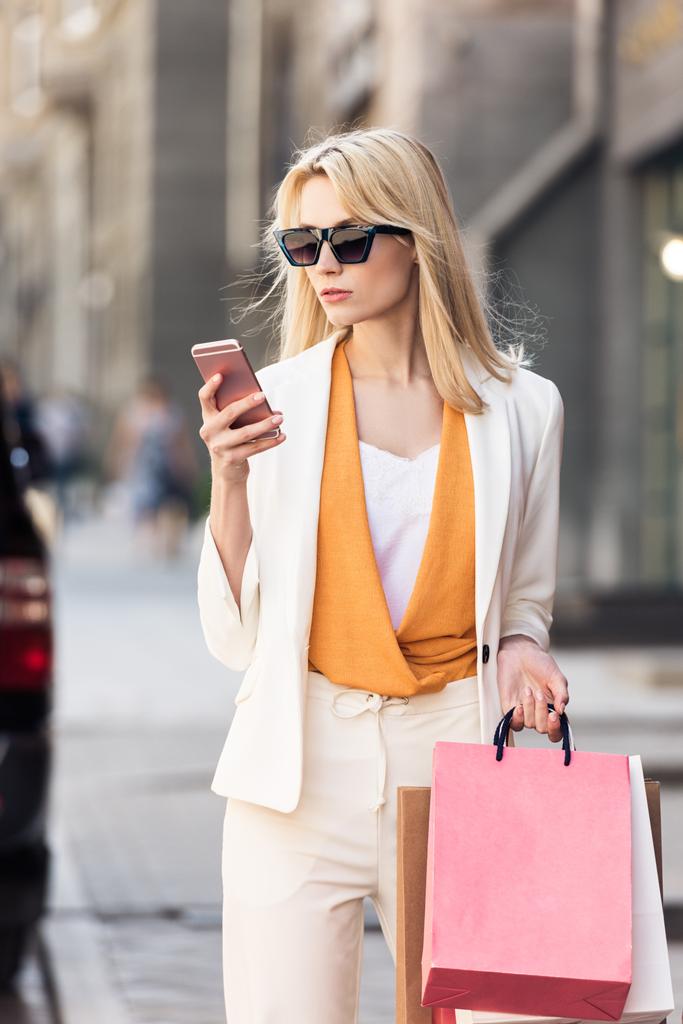 サングラスの買い物袋を押し、通りにスマート フォンを使用してスタイリッシュな金髪女性 - 写真・画像