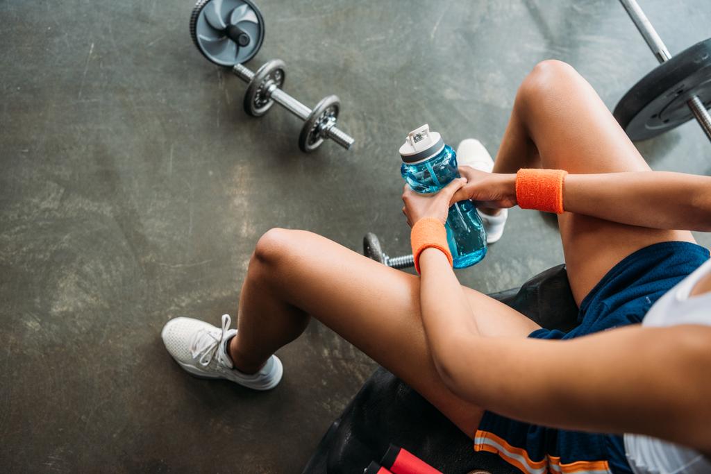 abgeschnittenes Bild einer Sportlerin in Armbändern, die mit einer Wasserflasche auf einem Trainingsreifen im Fitnessstudio sitzt  - Foto, Bild