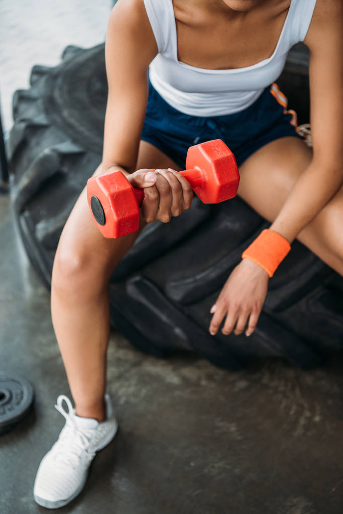 imagen recortada de la atleta haciendo ejercicio con la mancuerna y sentado en el neumático de entrenamiento en el gimnasio
  - Foto, imagen