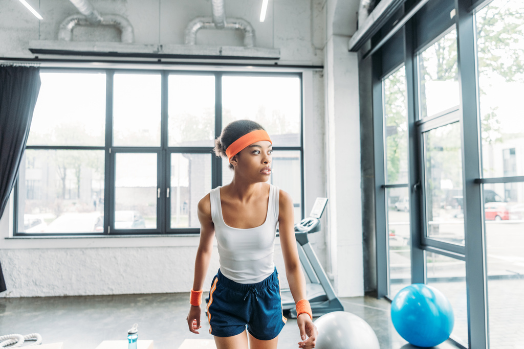 вид спереди на молодую африканскую спортсменку в повязке на голове и браслетах, тренирующуюся в спортзале
 - Фото, изображение