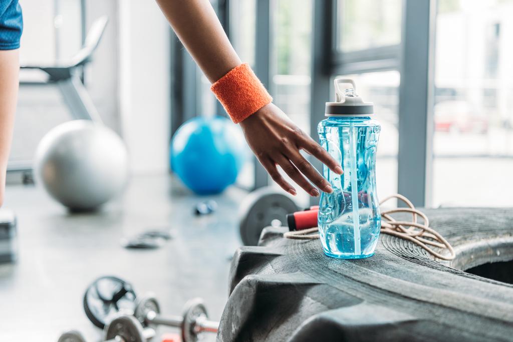 обрезанное изображение спортсменки в браслете, забирающей бутылку воды из тренировочной шины в спортзале
 - Фото, изображение