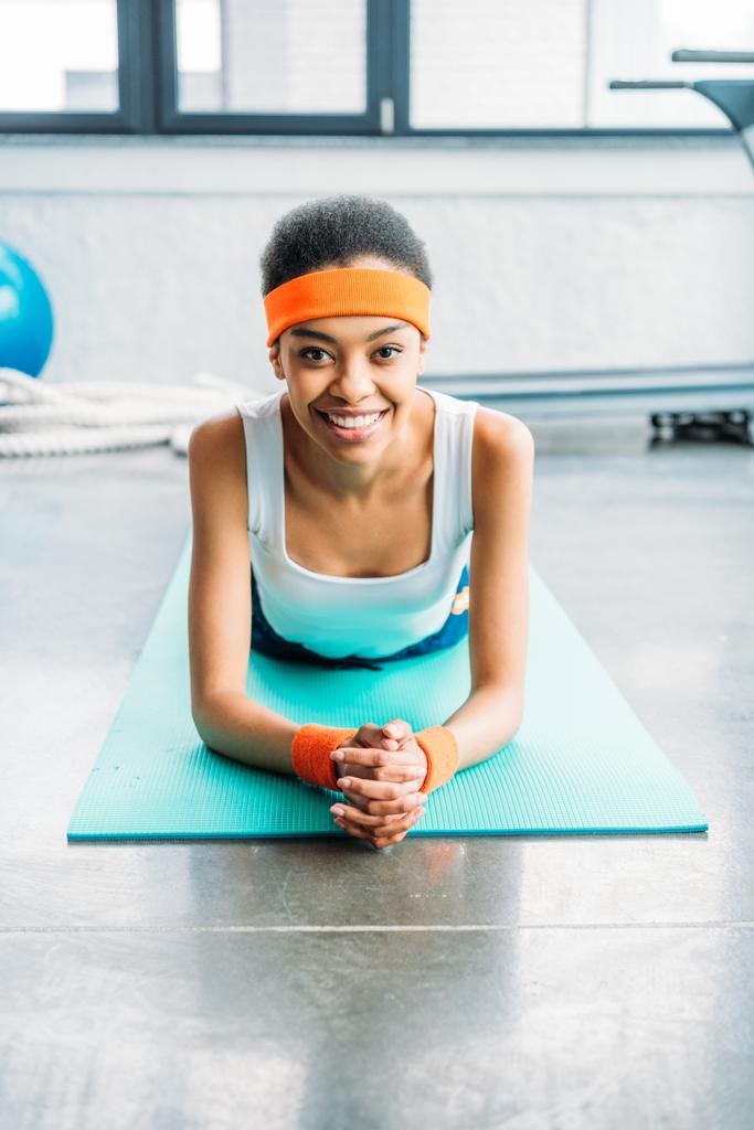 улыбающаяся африканская спортсменка в повязке на голове и браслетах, лежащих на фитнес-коврике в тренажерном зале
 - Фото, изображение