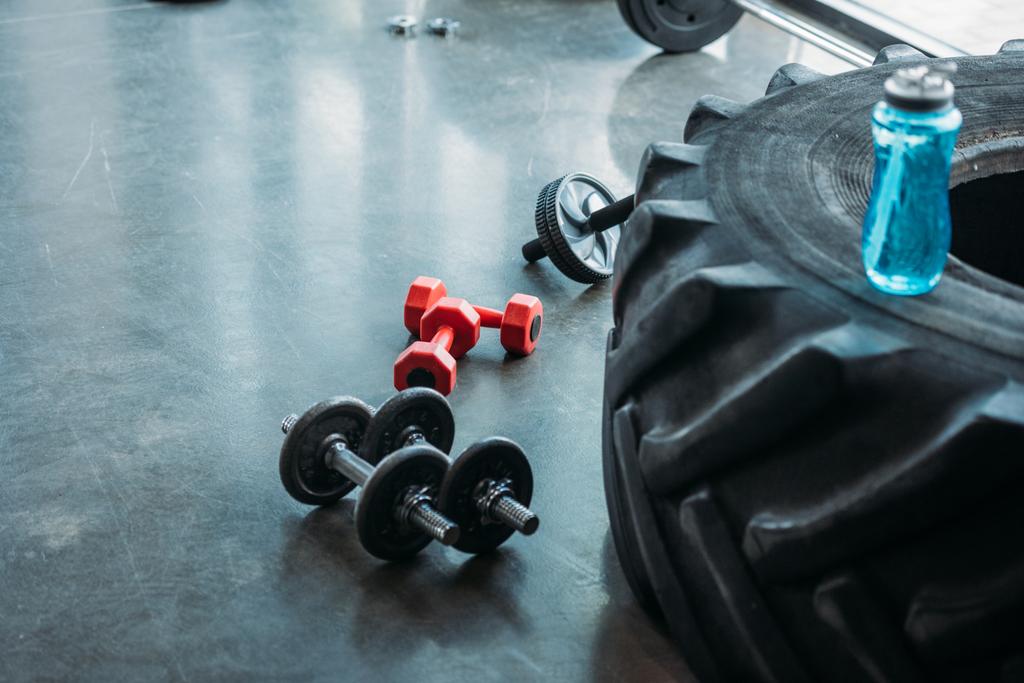 different dumbbells, abs roller and bottle of water on training tire at gym - Photo, Image