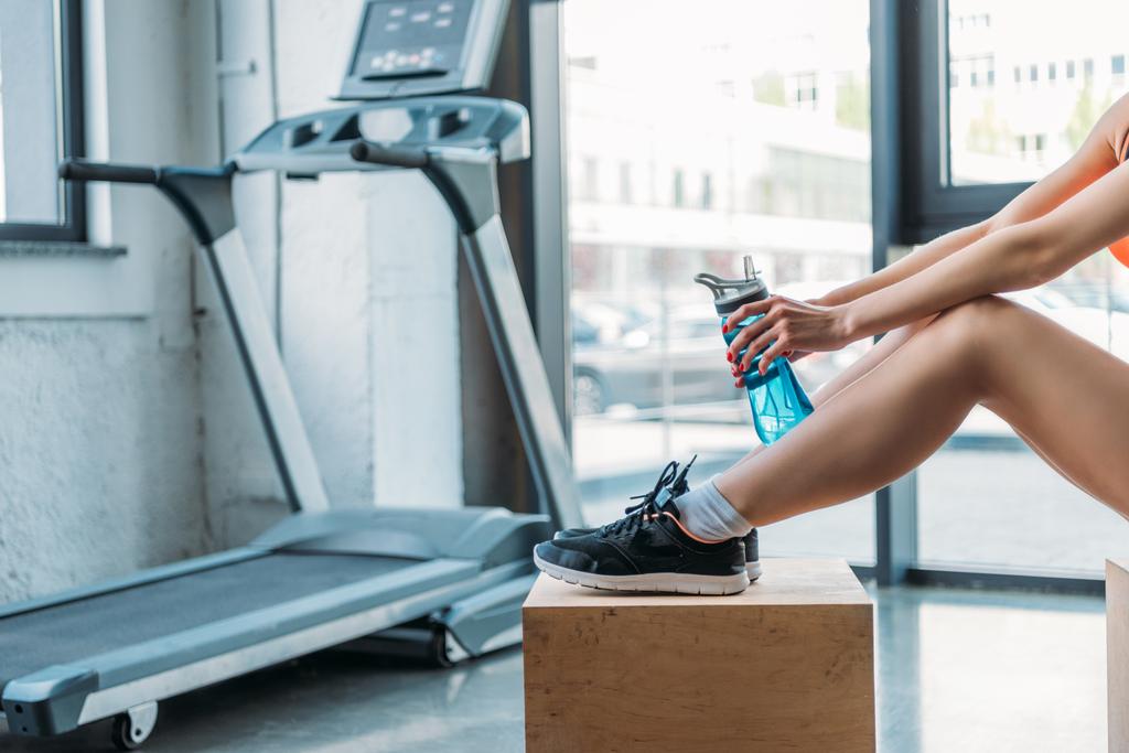 Oříznout záběr sportovkyně sedí na dřevěný box s lahví vody před běžícím pásu v posilovně - Fotografie, Obrázek