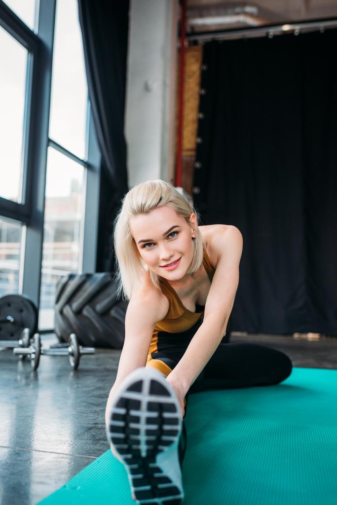 lachende atletische jongedame uitrekken op fitness mat op sportschool - Foto, afbeelding