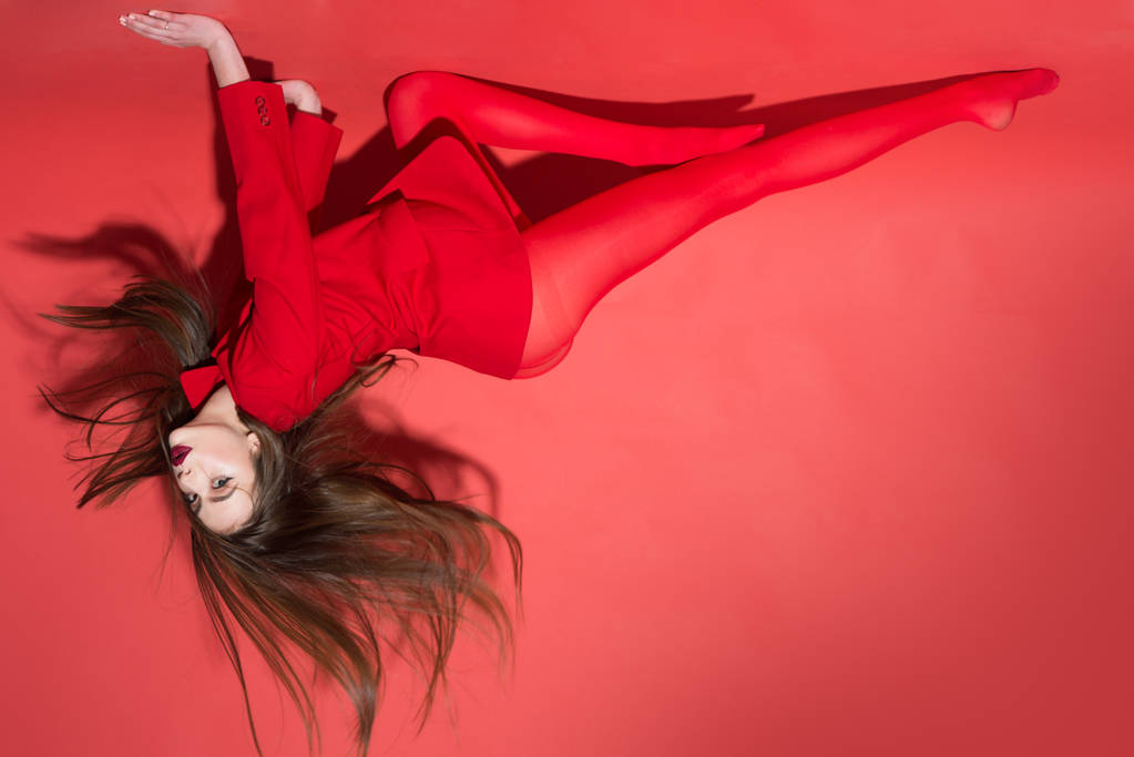 Beautiful female with stylish make-up and fluttering hair crawling on walls, wildness and energy of youth. Brunette girl in scarlet tights and jacket isolated on red background. - Photo, Image