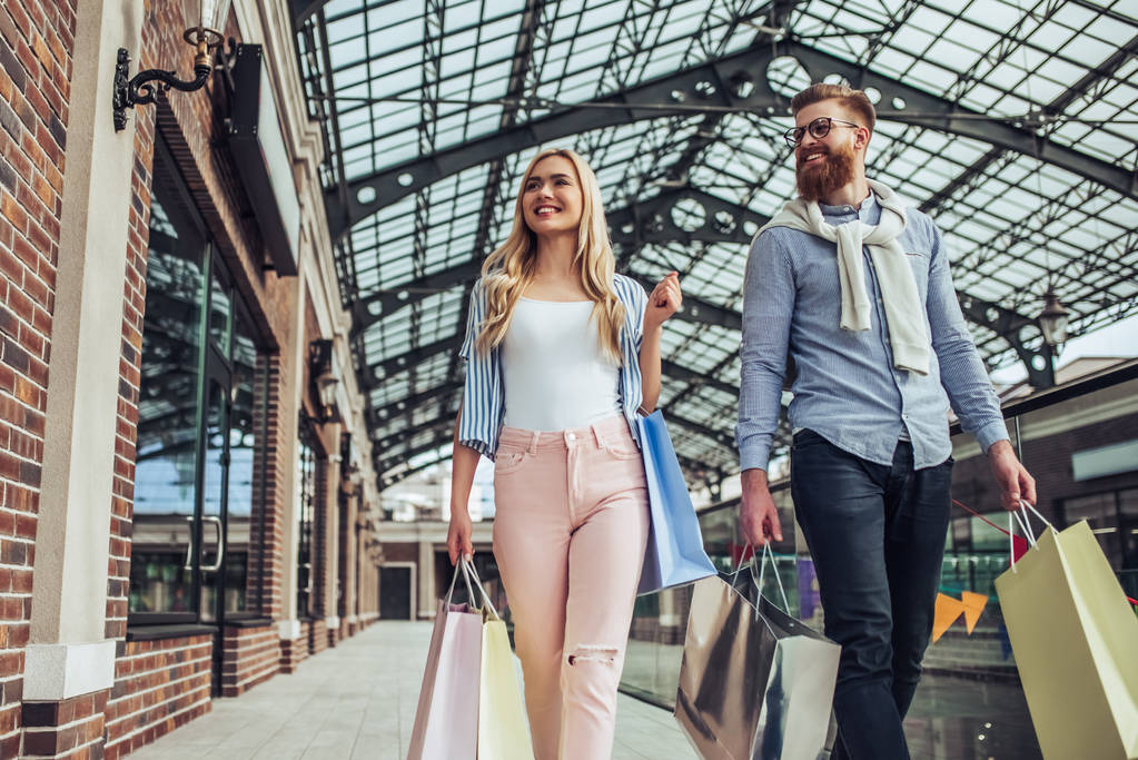 Stylish young couple is spending time together outdoors. Happy couple  is walking with shopping bags and doing shopping in the city. Attractive woman and handsome bearded man are having fun together. - Photo, Image
