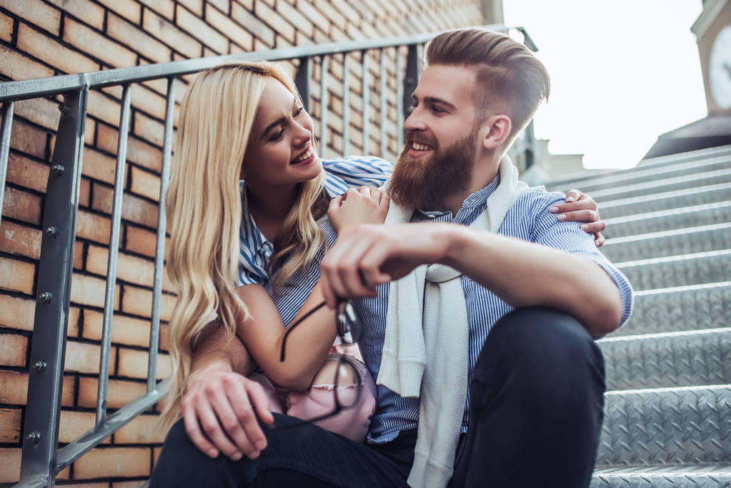Молода стильна щаслива пара проводить час разом на відкритому повітрі. Приваблива жінка і красивий бородатий чоловік сидять на сходах міста
. - Фото, зображення
