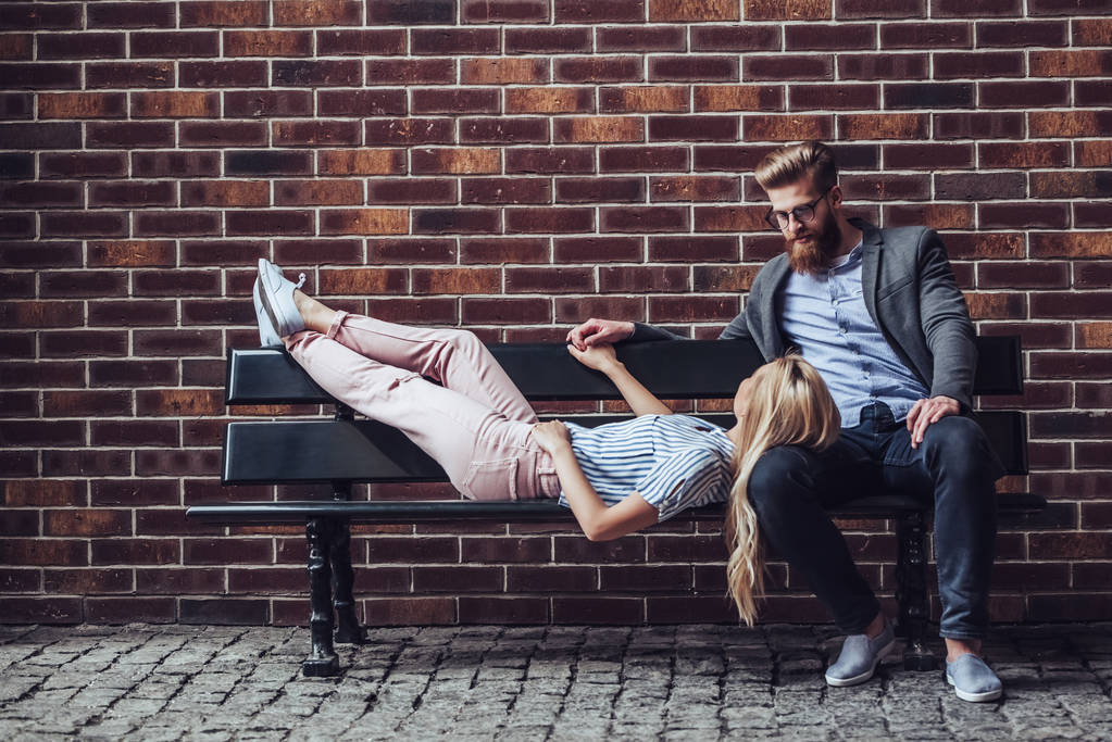Elegáns fiatal pár kiadási időt együtt szabadban. Vonzó nő és szép szakállas ember ül egy padon a tégla a háttérben a város. - Fotó, kép
