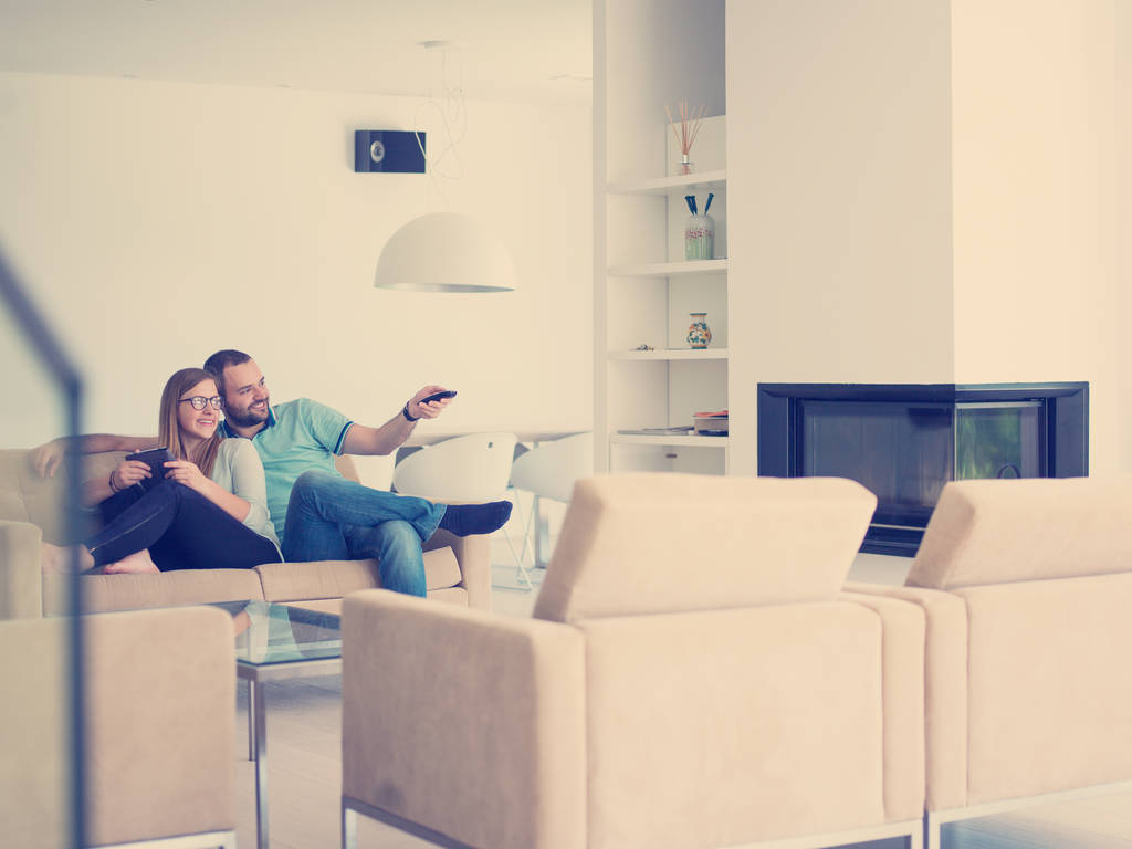 Молодая пара отдыхает на диване в роскошной гостиной, используя планшет и пульт дистанционного управления
 - Фото, изображение