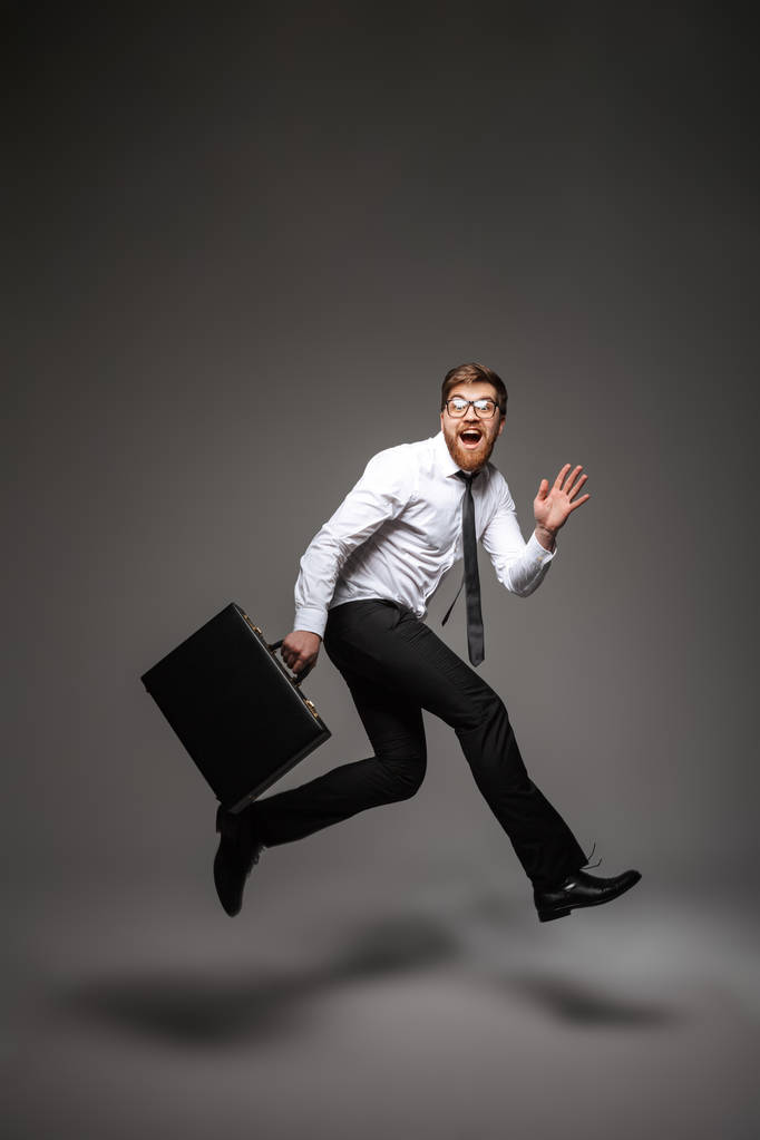 Полный портрет веселого молодого бизнесмена, позирующего, держа портфель и прыгая на сером фоне
 - Фото, изображение