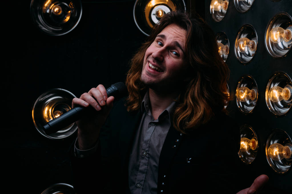Brutal singer with microphone on the stage, with long hair, against the background of bright lanterns, scene - Photo, Image