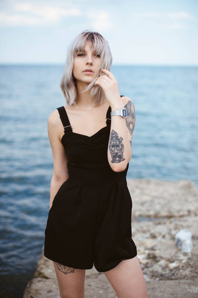 Hermosa mujer joven con tatuaje y vestido negro posando sobre fondo de mar
 - Foto, imagen
