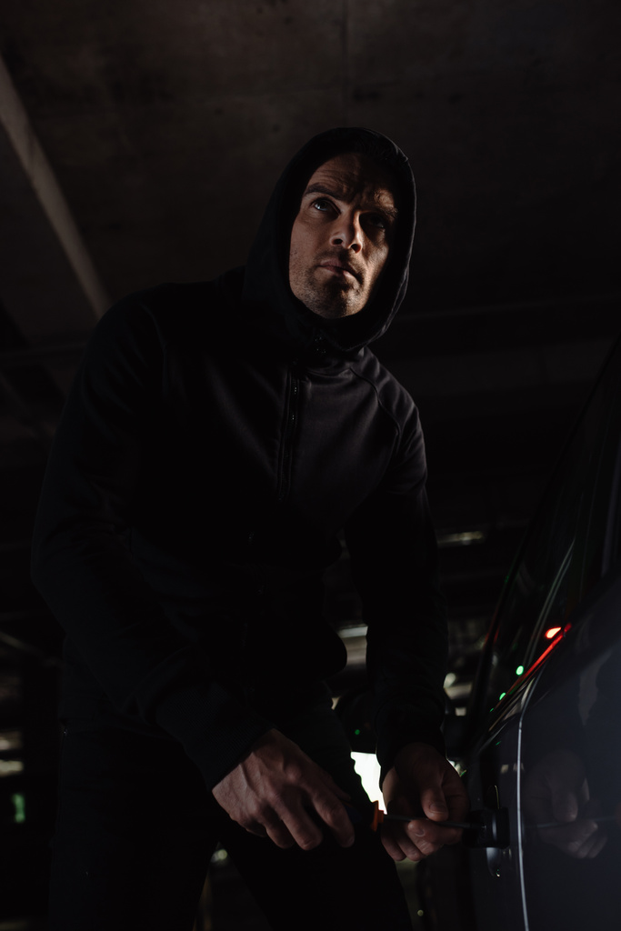 Männlicher Räuber in schwarzem Kapuzenpullover dringt mit Schraubenzieher in Auto ein  - Foto, Bild