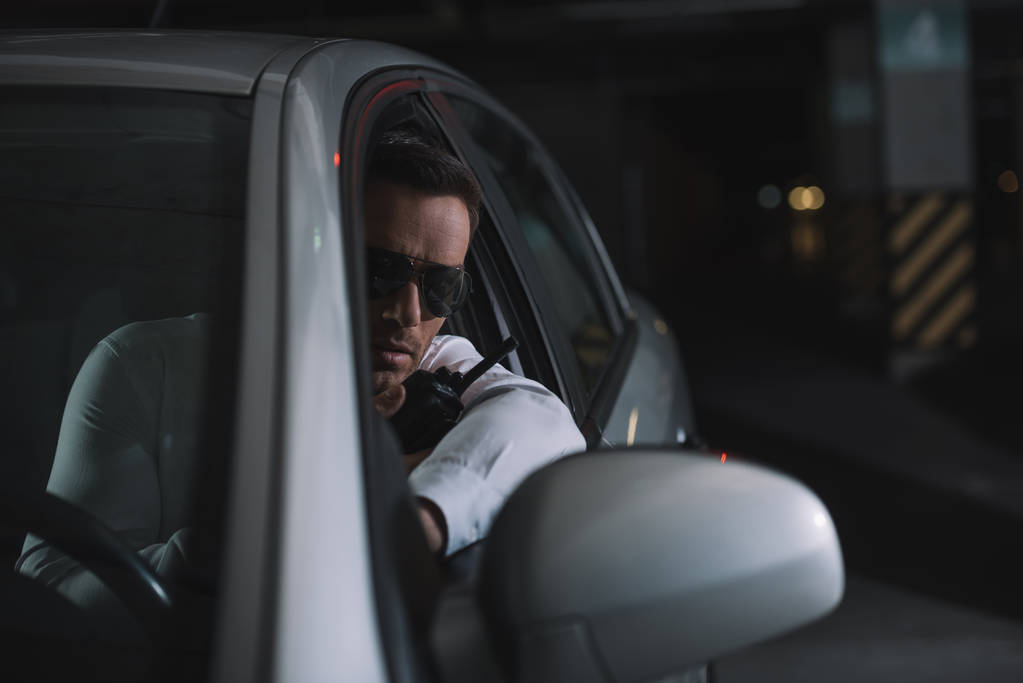 vakava mies soluttautunut agentti aurinkolaseissa tekemässä valvontaa ja käyttämällä talkie-radiopuhelinta autossa
 - Valokuva, kuva