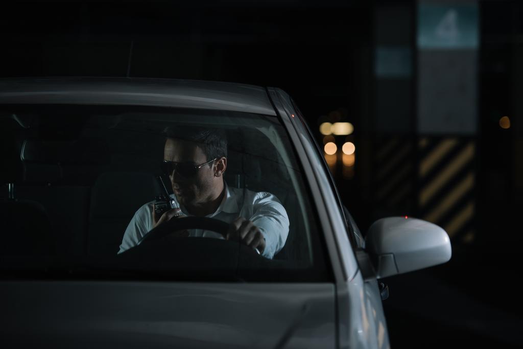 Vorderansicht eines männlichen Undercover-Agenten mit Sonnenbrille, der Überwachung betreibt und Talkie-Walkie im Auto benutzt - Foto, Bild