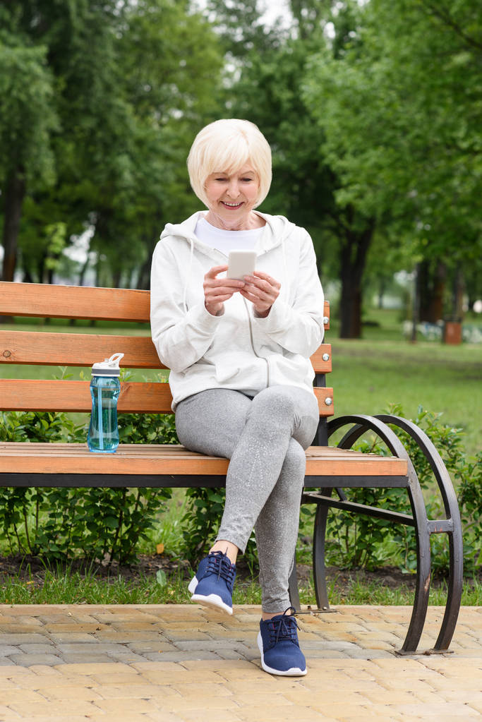 femme âgée en vêtements de sport à l'aide d'un smartphone assis sur un banc avec bouteille d'eau de sport
 - Photo, image