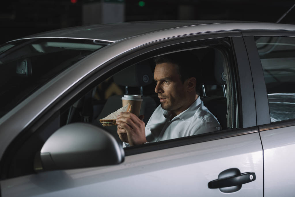 vista lateral del detective privado masculino bebiendo café y comiendo sándwich en su coche
 - Foto, imagen