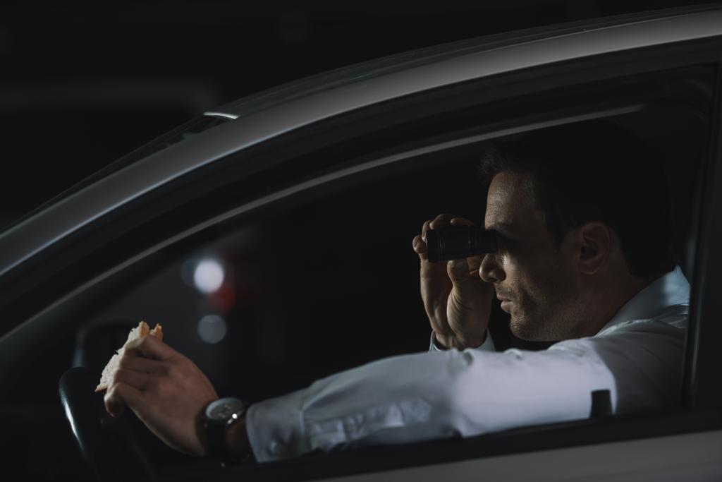 widok boczny tajny agent mężczyzna robi nadzoru przez lornetkę i Jedzenie Kanapki w samochodzie - Zdjęcie, obraz