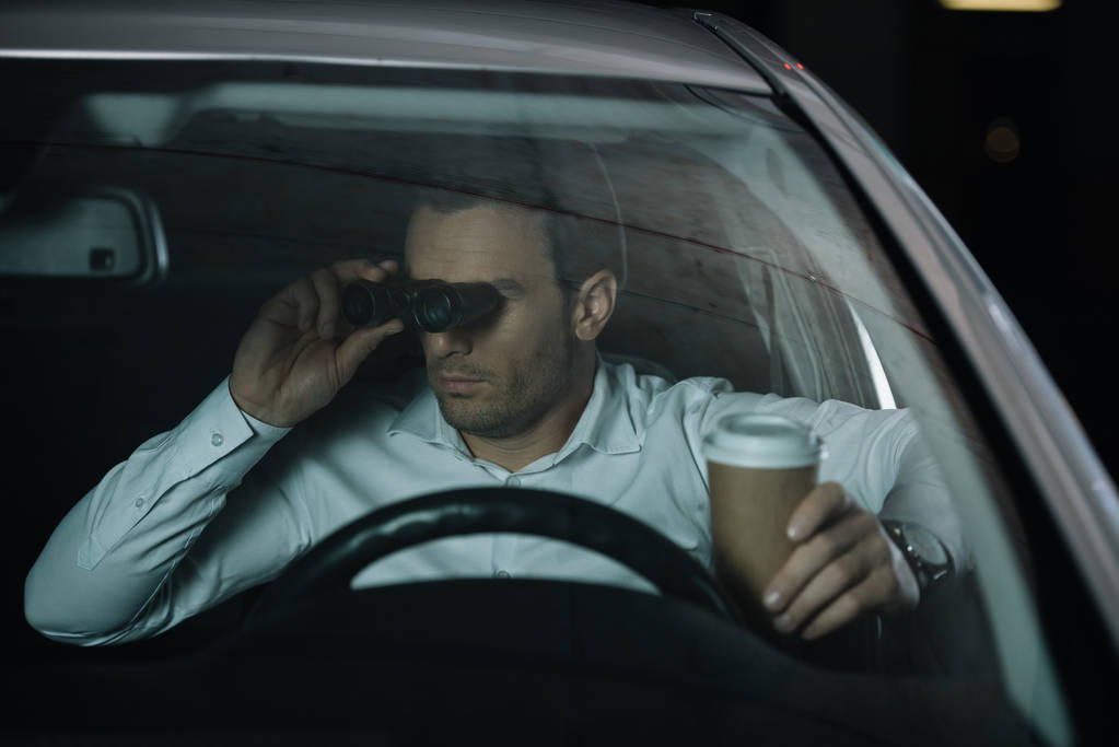 undercover male agent doing surveillance by binoculars and drinking coffee in car - Photo, Image