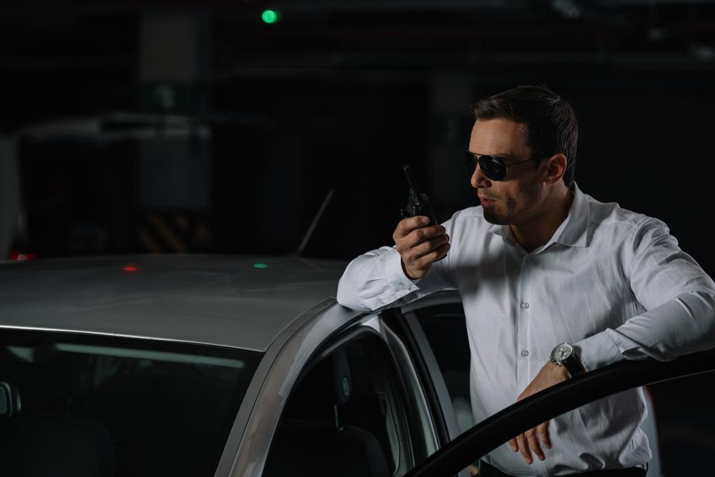 agente encubierto masculino en gafas de sol usando walkie talkie cerca del coche
  - Foto, Imagen