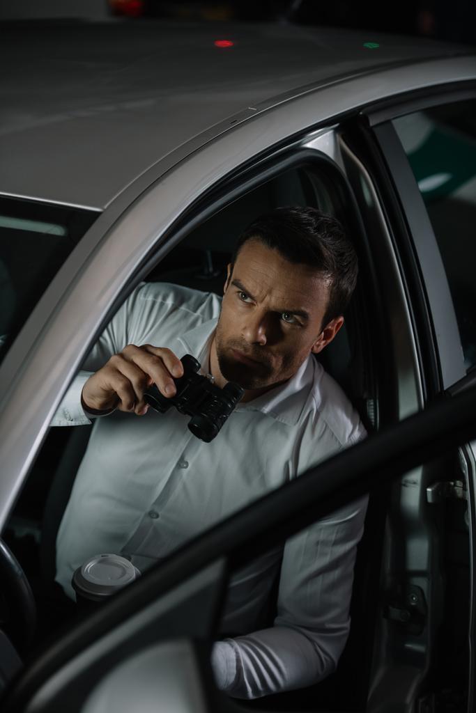 высокий угол зрения мужчины частного детектива с чашкой кофе делает наблюдение биноклем из автомобиля
 - Фото, изображение