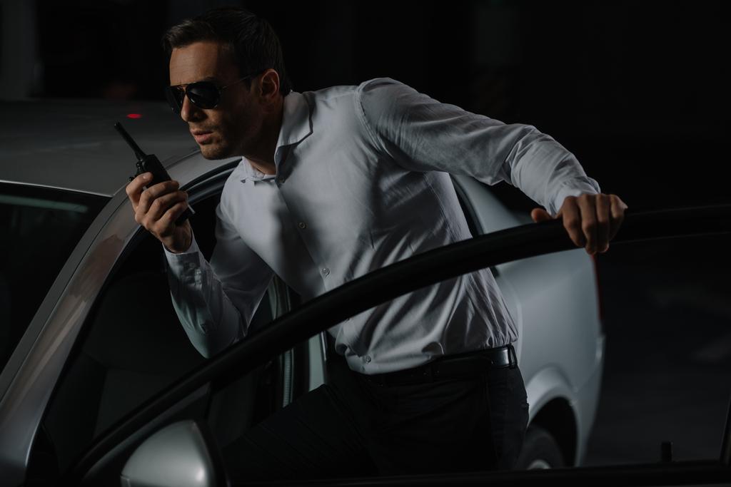 agente disfarçado masculino em óculos de sol usando walkie talkie perto do carro
  - Foto, Imagem