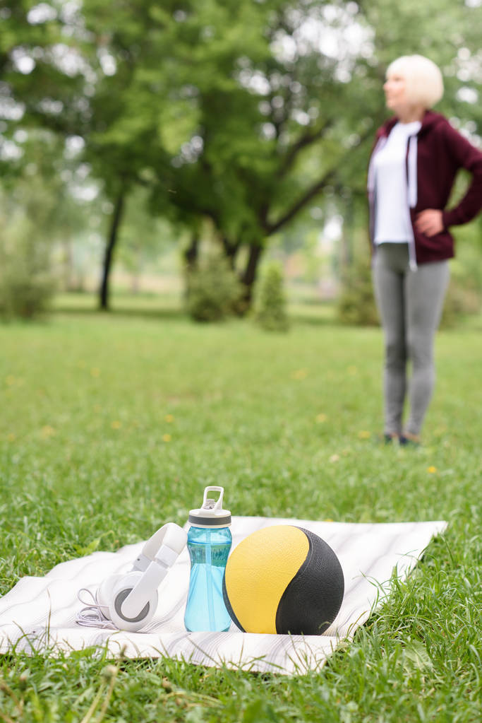 focus selettivo della donna nel parco con palla medica, acqua e cuffie sul tappetino yoga in primo piano
 - Foto, immagini