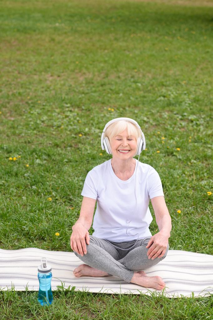 счастливая пожилая женщина слушает музыку в наушниках, сидя на коврике для йоги в парке
  - Фото, изображение