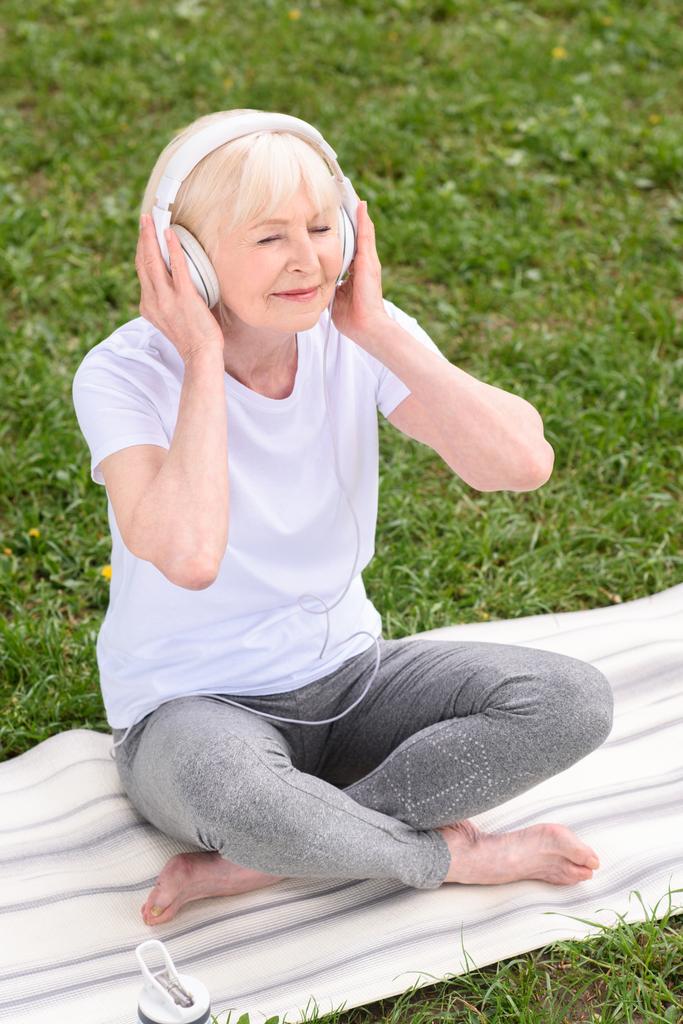kadın kıdemli yoga mat üzerinde otururken kulaklık ile müzik dinleme - Fotoğraf, Görsel