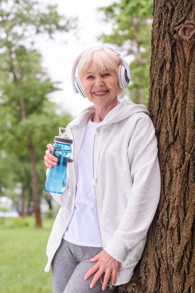 senior Sportvrouw glimlachend met sport fles luisteren van muziek met koptelefoon in park - Foto, afbeelding