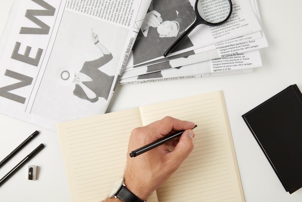 vista superior parcial de la persona que escribe en cuaderno en blanco, periódicos, lupa, cuaderno y material de oficina en gris
 - Foto, Imagen