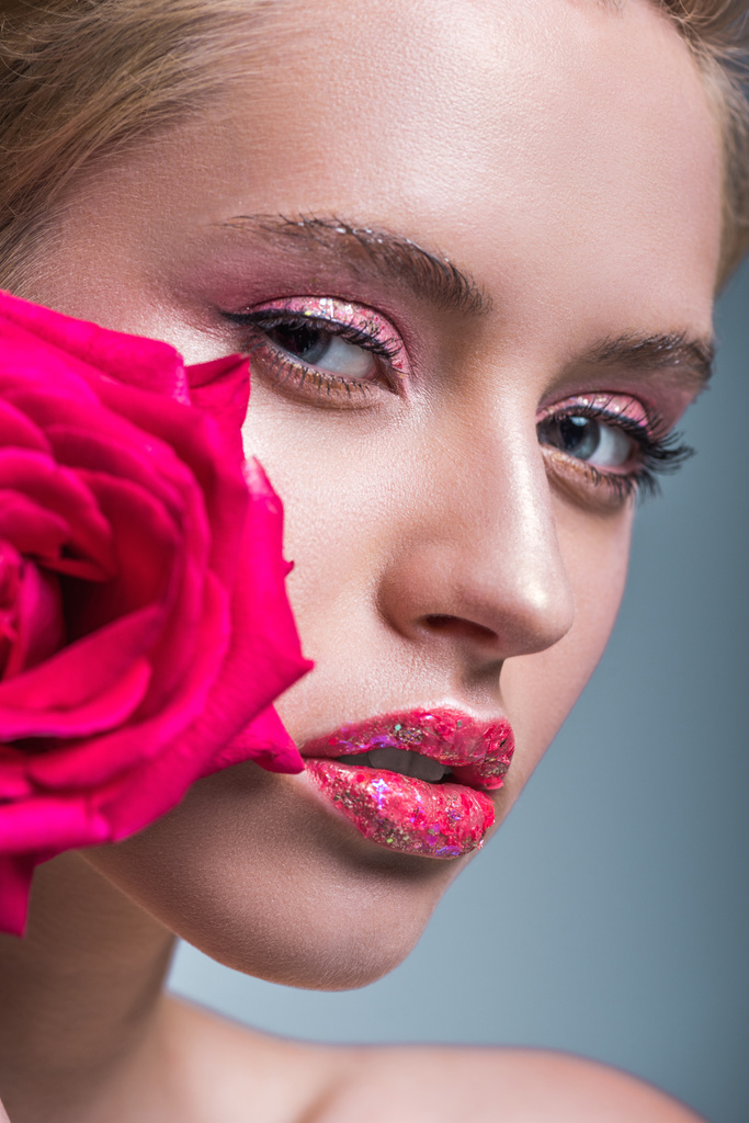 ελκυστική γυναίκα με glitter στα χείλη, κρατώντας κόκκινο τριαντάφυλλο και βλέπουν τα φωτογραφικών μηχανών που απομονώνονται σε γκρι - Φωτογραφία, εικόνα