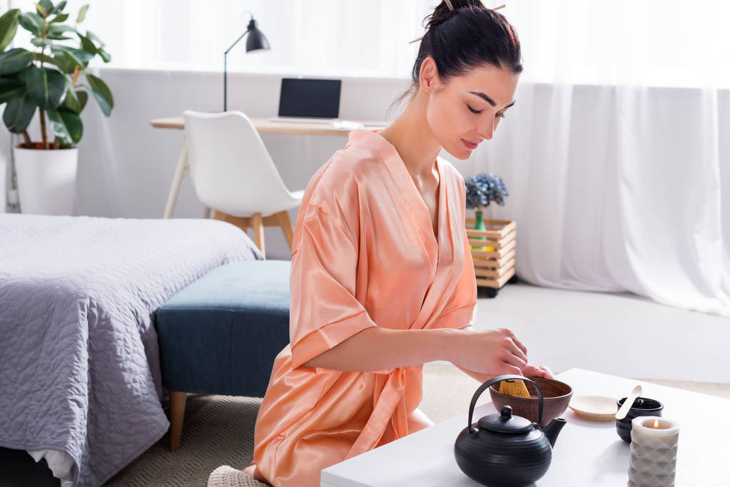привлекательная женщина в шелковом халате делает чай с венчиком во время чайной церемонии утром дома
 - Фото, изображение