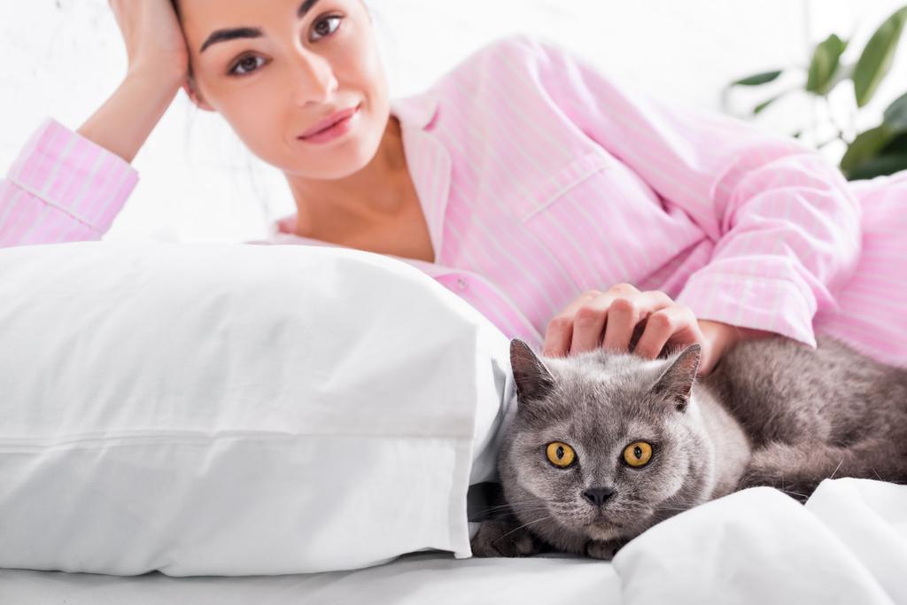 portrait of attractive woman in pajamas with britain shorthair cat resting on bed at home - Photo, Image