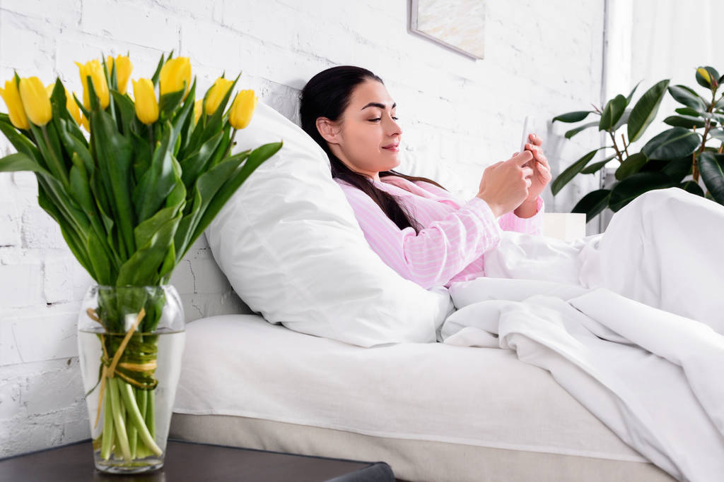Zijaanzicht van aantrekkelijke vrouw in pyjama met behulp van de smartphone tijdens het rusten op bed thuis - Foto, afbeelding