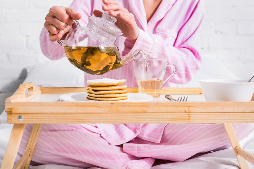 Teilbild einer Frau im rosafarbenen Pyjama, die grünen Tee in Tasse gießt, während sie zu Hause im Bett frühstückt - Foto, Bild