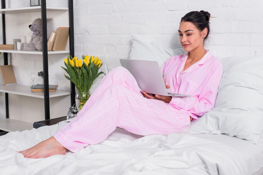 Улыбающаяся женщина в пижаме с ноутбуком в постели дома
 - Фото, изображение