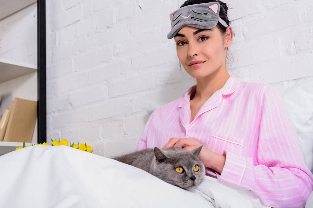 Πορτραίτο γυναίκας στο πιτζάμες και μάσκα ύπνου με μεγάλη Βρετανία shorthair γάτα ανάπαυση στο κρεβάτι στο σπίτι - Φωτογραφία, εικόνα