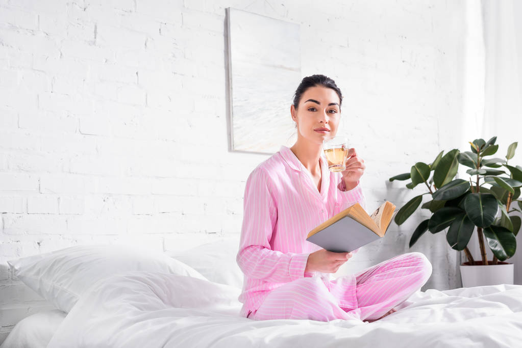 Kobieta w piżamie z kubkiem gorącej herbaty i siedzi na łóżku rano w domu książki - Zdjęcie, obraz